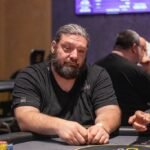 Ein Mann und eine Frau spielen Poker an einem Tisch in einem eleganten Casino. Der Mann hat eine Hand mit Karten in der Luft, während die Frau konzentriert ihre Karten betrachtet. Im Hintergrund sind weitere Spieler