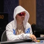 Ein Mann und eine Frau sitzen an einem Pokertisch und spielen Poker. Der Mann hat eine Hand mit Karten in der Hand, während die Frau konzentriert auf ihre Karten schaut. Im Hintergrund sind Pokerchips
