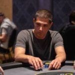 Ein Mann sitzt an einem Pokertisch und hält zwei Karten in der Hand, während er konzentriert auf die Gemeinschaftskarten schaut. Andere Spieler sind im Hintergrund zu sehen.