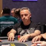 Ein Mann sitzt an einem Pokertisch und hält zwei Spielkarten in der Hand. Er schaut konzentriert auf den Tisch, auf dem Pokerchips und Karten liegen. Im Hintergrund sind weitere Spieler