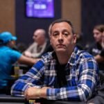 Ein Mann sitzt an einem Pokertisch und betrachtet konzentriert seine Karten. Im Hintergrund sind weitere Spieler zu sehen. Die Atmosphäre ist gespannt und fokussiert.