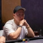 Ein glücklicher Mann sitzt an einem Pokertisch und hält eine Hand mit Spielkarten. Er lächelt und hat eine entspannte Haltung. Im Hintergrund sind andere Spieler und Pokerchips zu sehen.