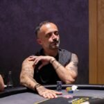 Ein Mann spielt Poker an einem Tisch in einem eleganten Casino. Er hält Karten in der Hand und setzt Chips. Andere Spieler und ein Dealer sind im Hintergrund zu sehen.