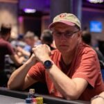 Ein Mann spielt Poker an einem Tisch mit grüner Filzdecke, umgeben von anderen Spielern. Er hält Karten in der Hand und betrachtet sie konzentriert. In der Mitte des Tisches