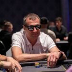 Ein Mann sitzt an einem Pokertisch und hält zwei Karten in der Hand, während er ernst und konzentriert auf den Tisch starrt. Andere Pokerspieler sind im Hintergrund verschwommen