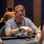 Ein grüner Pokertisch mit Spielkarten, Pokerchips und Getränken; Menschen in der Nähe, die eine Runde Poker spielen.
