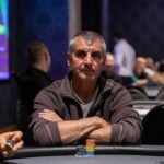 Ein Mann in einem eleganten Anzug sitzt an einem Pokertisch und hält eine Hand mit Spielkarten. Er schaut konzentriert auf seine Karten, während andere Spieler im Hintergrund zu sehen sind. Die Atmos