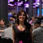 Ein Mann in einem schwarzen Anzug und einer roten Krawatte sitzt an einem Pokertisch und hält seine Spielkarten in der Hand. Im Hintergrund sind Chips und weitere Spieler zu sehen.