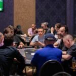 Ein Mann und eine Frau sitzen an einem Pokertisch und spielen eine Partie Poker. Der Mann hält Karten in der Hand und schaut konzentriert auf den Tisch, während die Frau mit ernstem Gesichtsaus