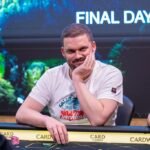 Ein Mann in einem schwarzen Anzug sitzt an einem Pokertisch und hält seine Karten. Andere Spieler sind im Hintergrund zu sehen.