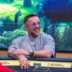 Ein Mann in einem schwarzen Anzug und einer roten Krawatte sitzt an einem Pokertisch und hält eine Hand mit Spielkarten. Um ihn herum sitzen vier weitere Personen, von denen man nur die Hände und