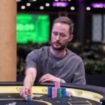 Ein Mann spielt Poker an einem Tisch in einem eleganten Casino. Er hat eine ernste Miene und hält Karten in der Hand, während er von anderen Personen umgeben ist. Die Atmosphäre ist gespannt und glamour