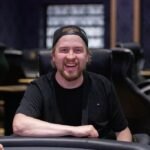 Ein Mann mit schwarzer Sonnenbrille und Kappe sitzt an einem Pokertisch und hält zwei Spielkarten in der Hand. In der Mitte des Tisches sind Pokerchips und weitere Karten zu sehen.