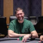 Ein Mann sitzt an einem Pokertisch und hält zwei Spielkarten in der Hand. Er trägt einen Anzug und eine dunkle Sonnenbrille. Im Hintergrund sind Pokerchips und andere Spieler zu sehen.