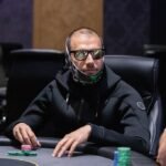 Ein eleganter Pokertisch mit grüner Filzoberfläche und mehreren Spielkarten und Chips darauf. Im Hintergrund sind Spieler in einer intensiven Pokerrunde zu sehen.