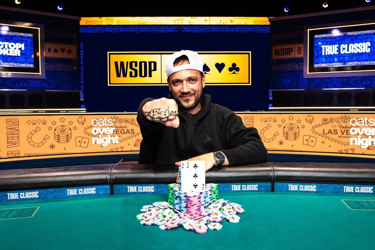 Ein Mann in einem dunklen Anzug und einer Krawatte sitzt lächelnd an einem Pokertisch und hält Spielkarten in der Hand.