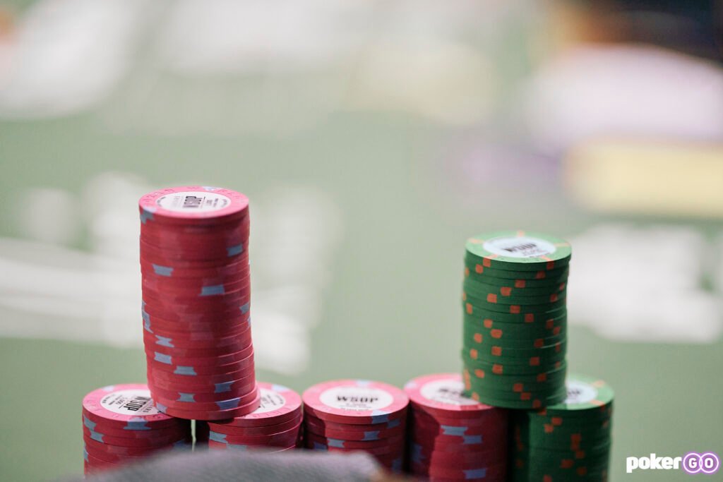 Ein Stapel von bunten Pokerchips auf einem grünen Tisch, der während der World Series of Poker platziert ist. Ein Spieler ist im Hintergrund zu sehen, der seine Karten betrachtet.