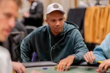 Ein Mann in einem schwarzen Anzug sitzt an einem Pokertisch während eines Turniers. Er hat einen konzentrierten Ausdruck und betrachtet seine Karten. Um ihn herum sind andere Spieler und Zuschauer zu