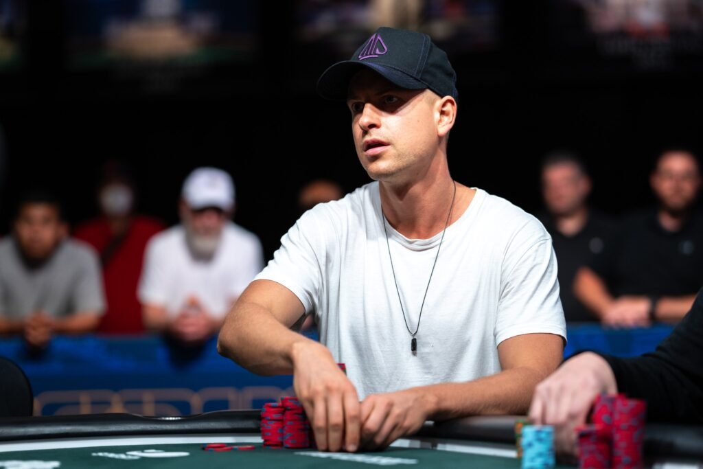 Ein junger Mann mit dunklen Haaren und einem Bart sitzt an einem Pokertisch und betrachtet konzentriert seine Karten. Er trägt ein dunkles Hemd und hat eine ernste Miene.